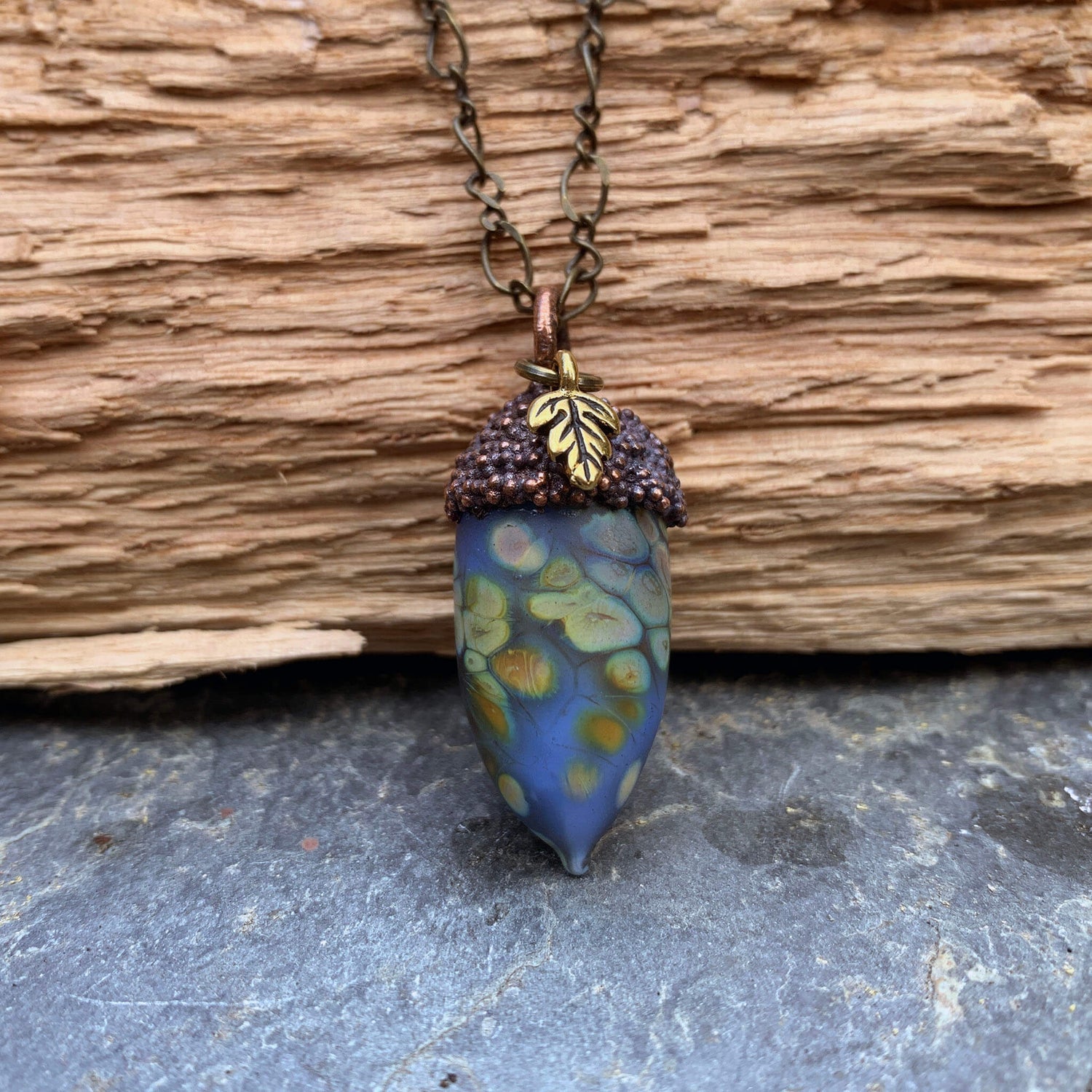 Limited Edition Glass and Copper Acorn Pendant in Periwinkle, photographed against wood on a stone surface.